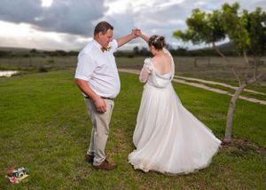 A couple dancing together outdoors on grass with storm clouds and trees in the background at sunset.