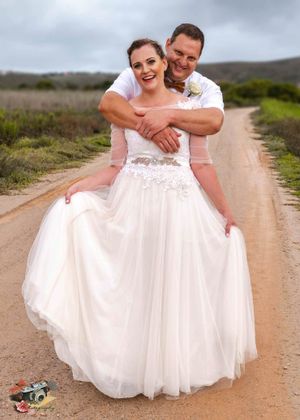 Wedding couple shares playful moment on country road with groom embracing bride in flowing white gown.