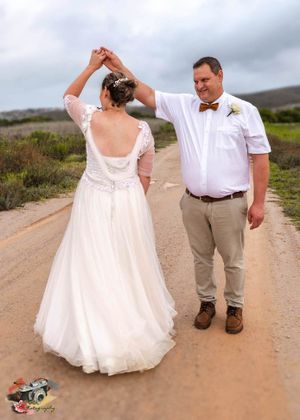 A bride and groom share a romantic dance pose on a dirt road with cloudy skies and rolling hills in the background.