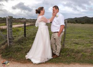 A rustic country wedding photo with couple laughing together on a dirt path near wooden fencing under cloudy skies.