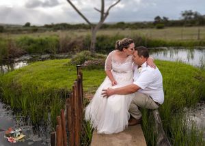 A romantic couple sits together on a rustic wooden bridge over a pond in a scenic country setting during a wedding celebration.