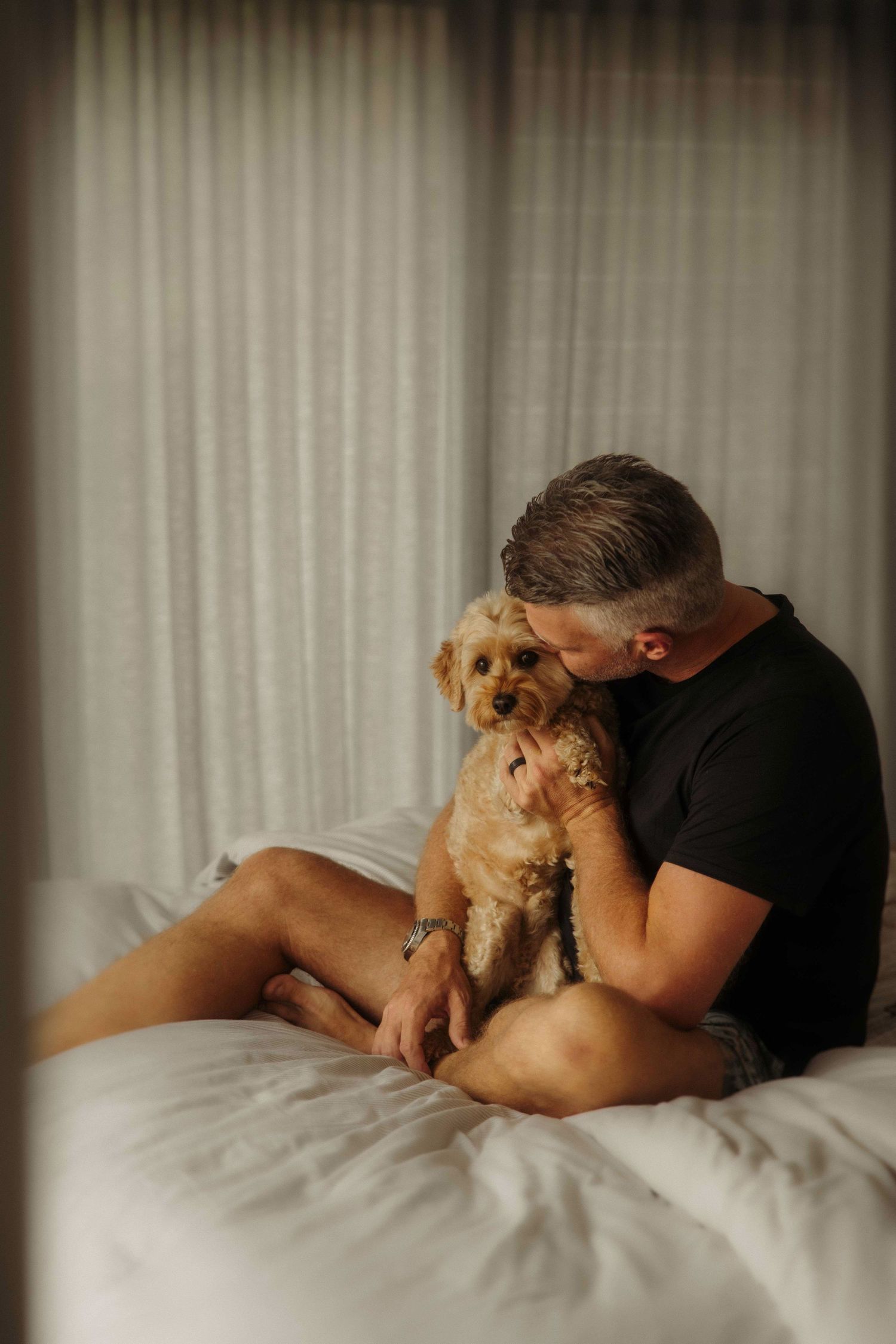 Intimate portrait of someone in black t-shirt cuddling with small brown dog on white bed.