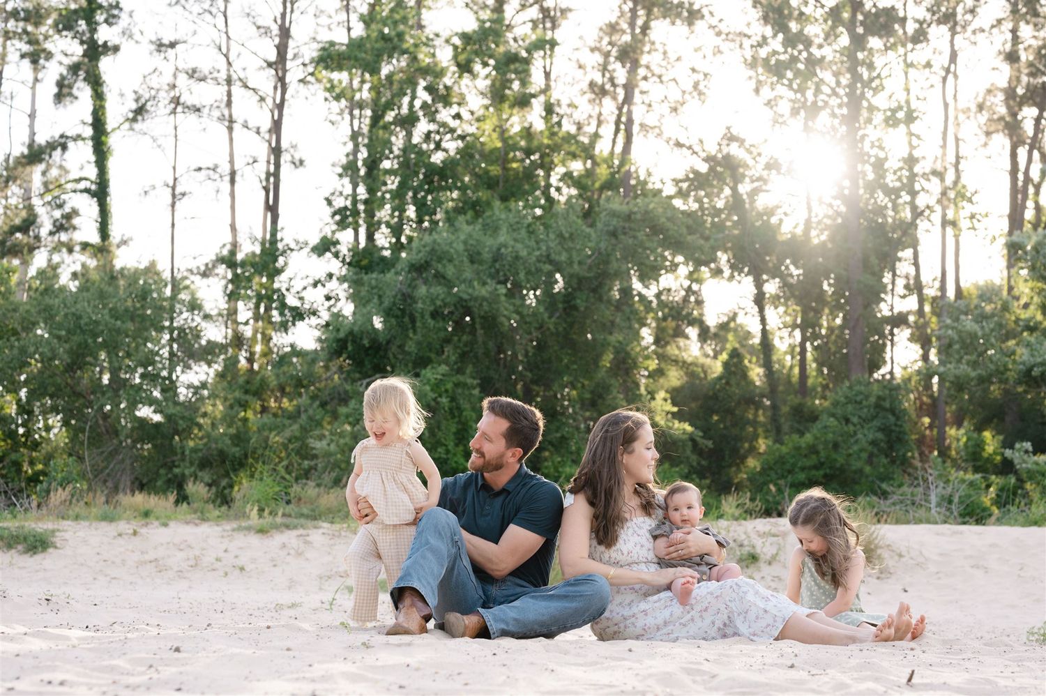 A group sits together on a sandy beach at sunset with tall pine trees in the background.