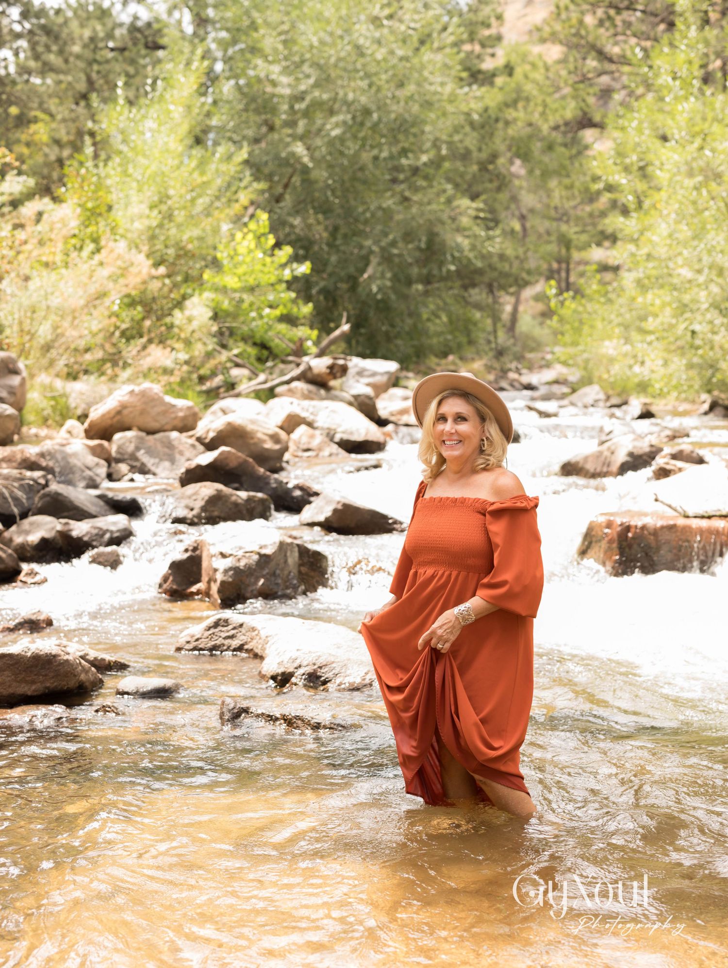 A person in a flowing rust-colored dress stands in a rocky mountain stream on a sunny summer day.