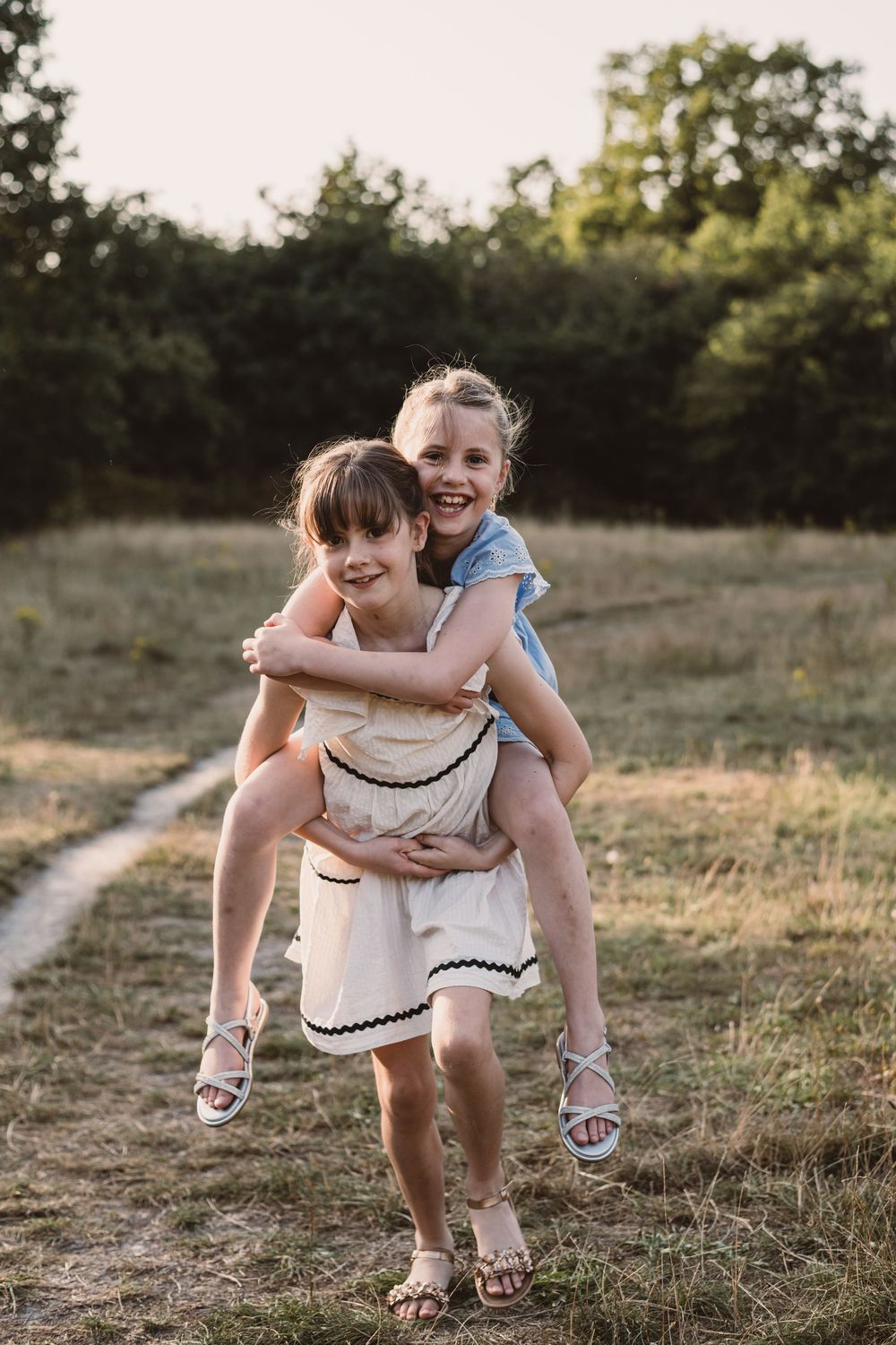 Two young children play piggy back on a grassy hillside path during golden hour.