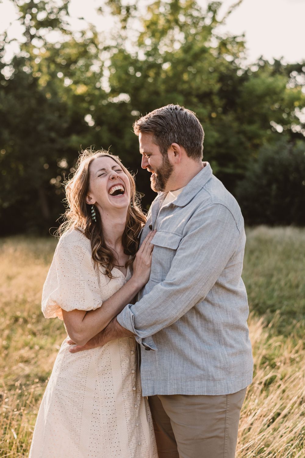A couple shares joyful laughter together in a sunlit field with tall grass during an outdoor photoshoot at golden hour.