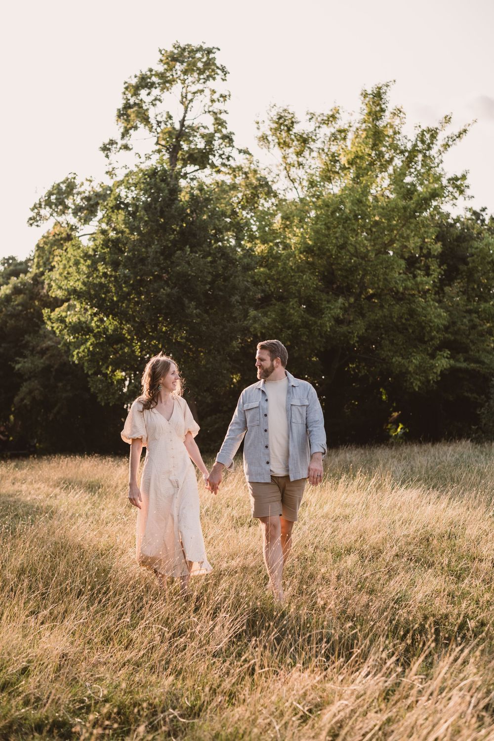 A couple walks hand in hand through a golden meadow at sunset with trees in the background.