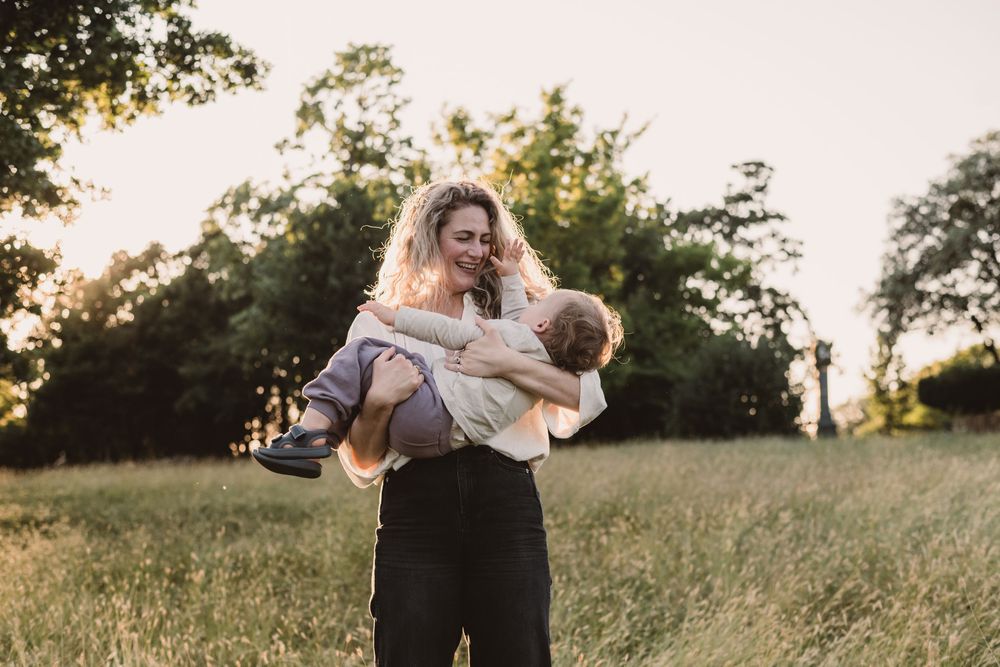 A parent holds a small child in a grassy field at sunset with trees silhouetted in the background.