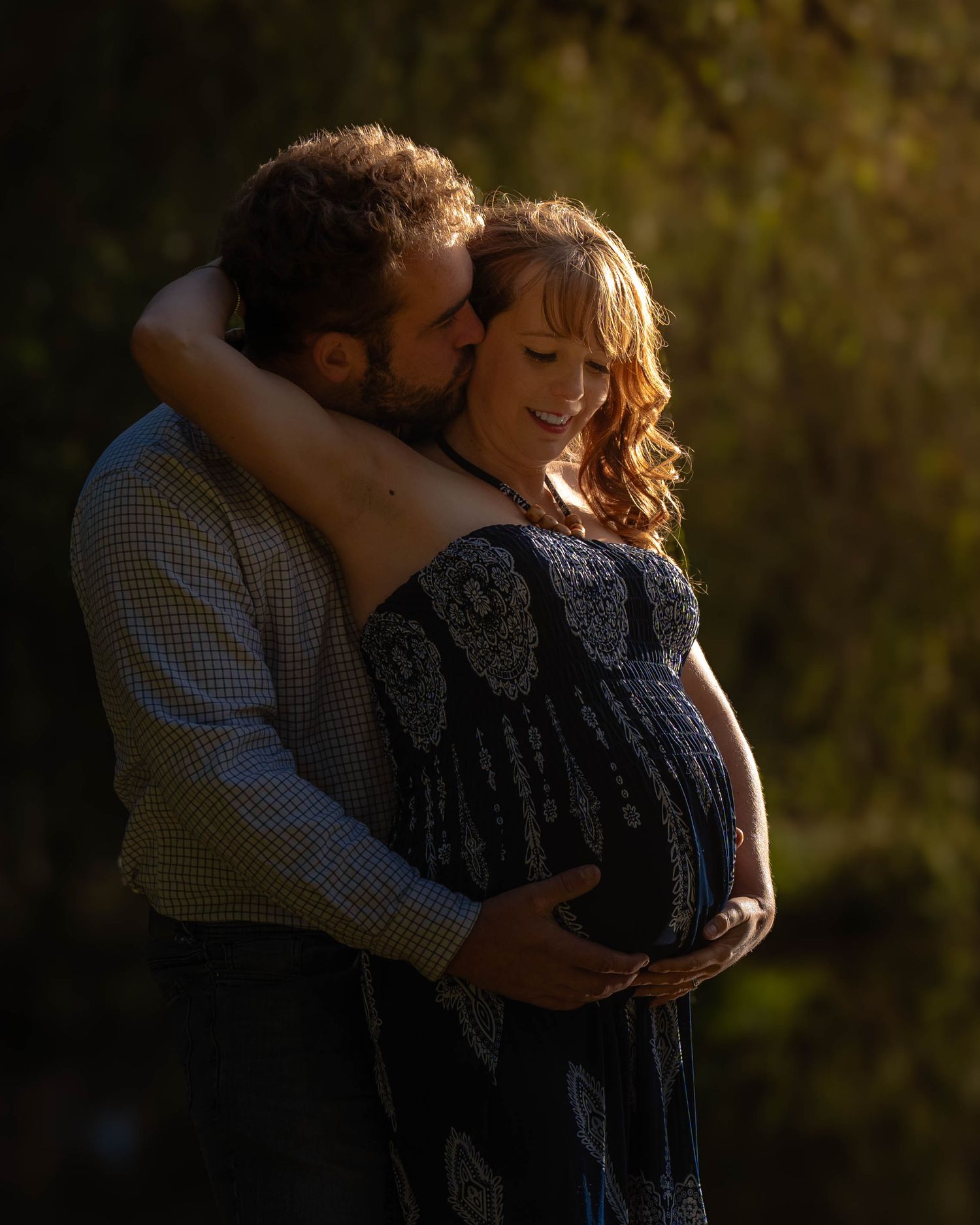 A silhouetted couple embraces in warm sunlight during a maternity photoshoot outdoors.