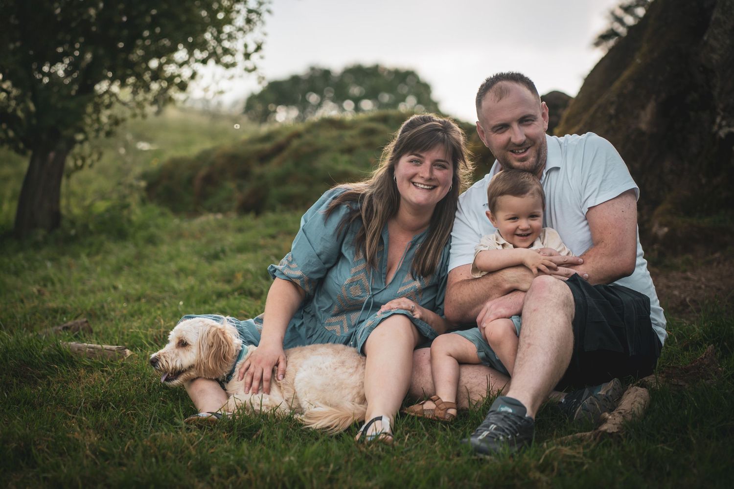 A family sits together on grass with their golden retriever dog during a casual outdoor photoshoot.