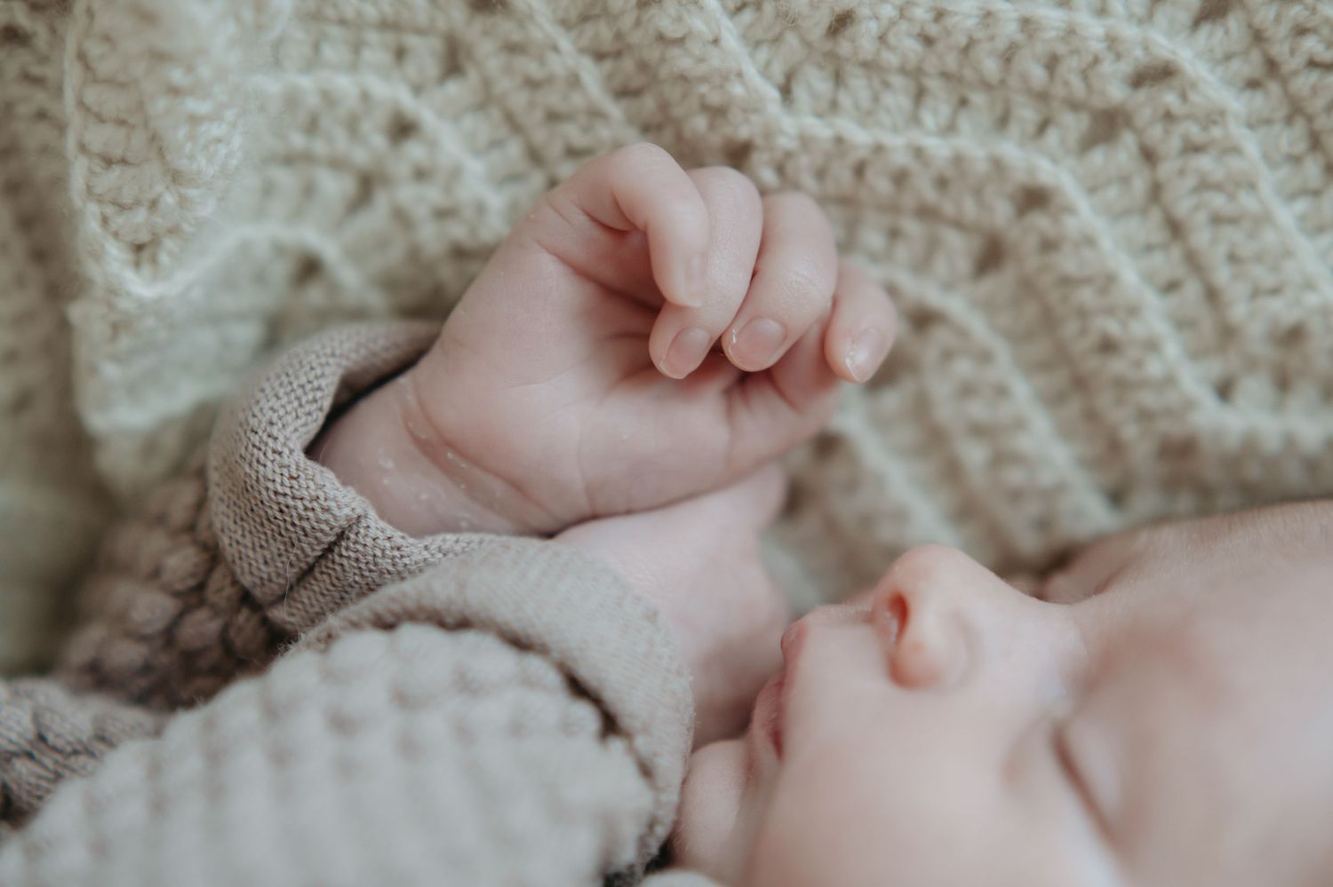 Close-up view of tiny sleeping baby's fist against soft knitted blanket in muted beige tones.