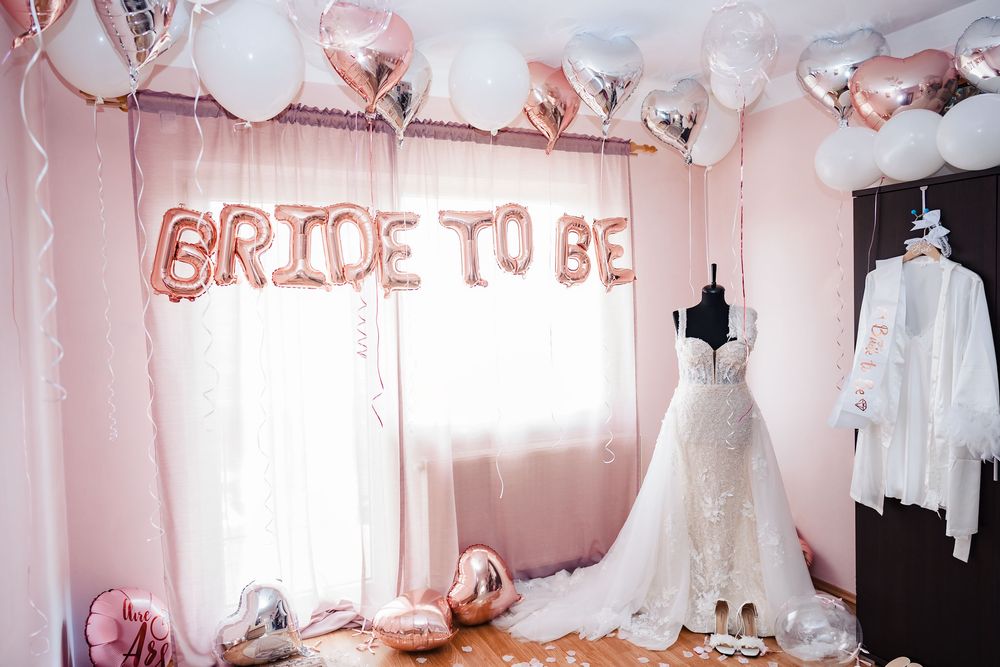 Wedding dress display with rose gold 'Bride to Be' balloon letters and pink decorations.
