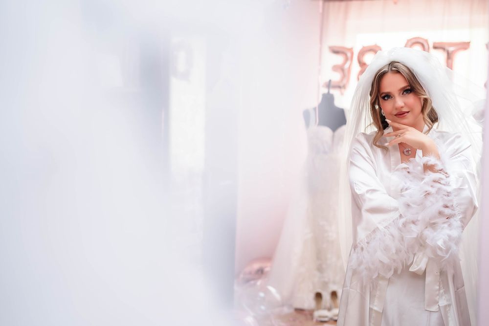 Bride in white silk robe poses thoughtfully in a softly lit dressing room.