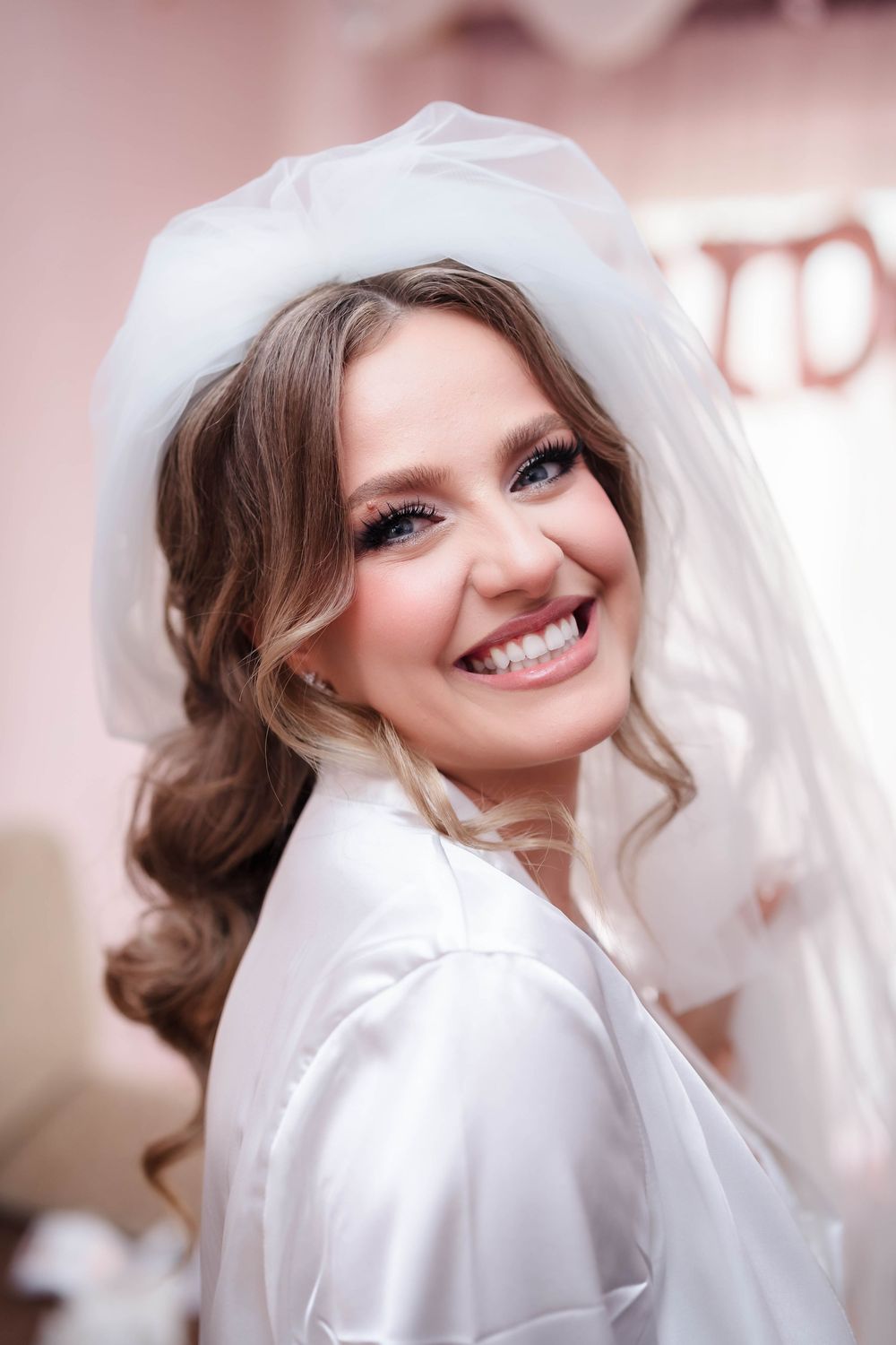 Bride wearing white robe and veil smiles radiantly during wedding preparations.