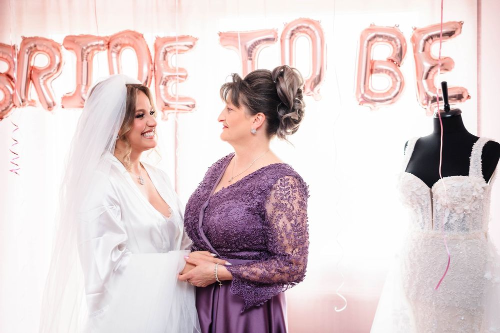 A bride in white dress shares a tender moment with a woman in purple lace gown against a pink backdrop with balloons.