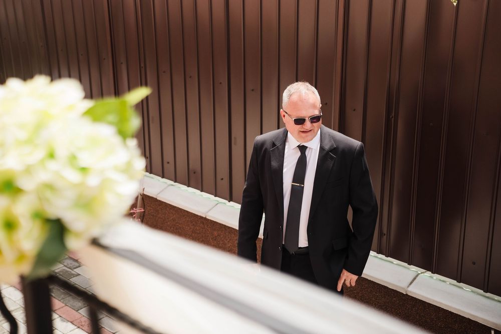 Man in dark suit and sunglasses stands near wooden paneled wall at formal event.