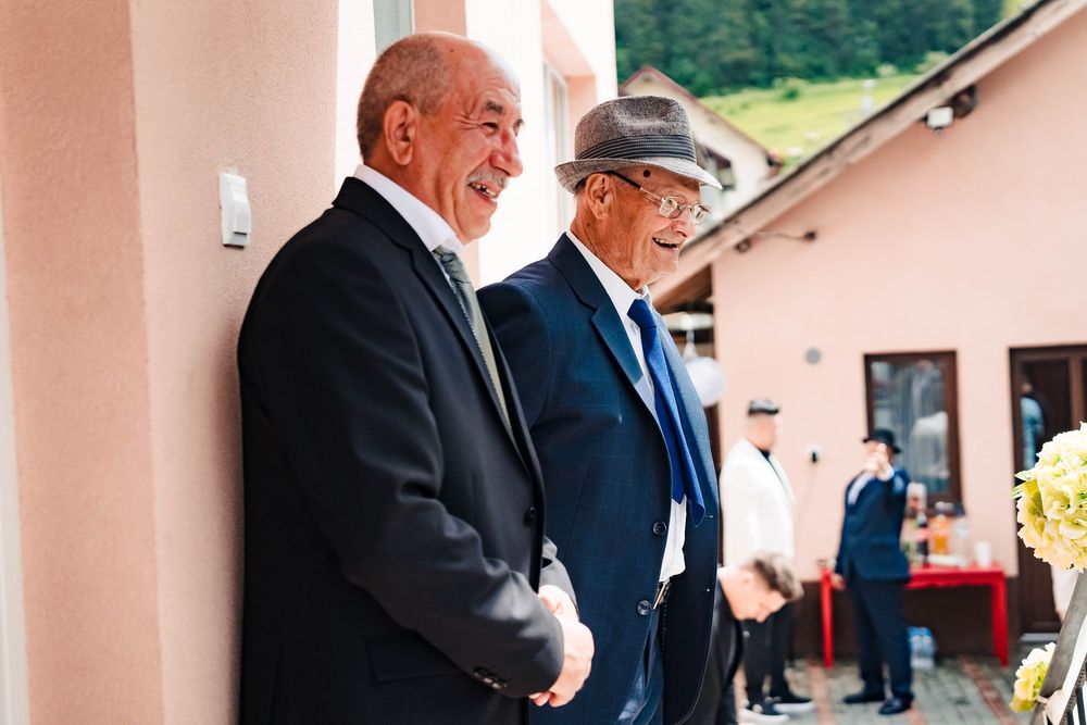 Two elderly men in formal attire share a moment outside a pink building.
