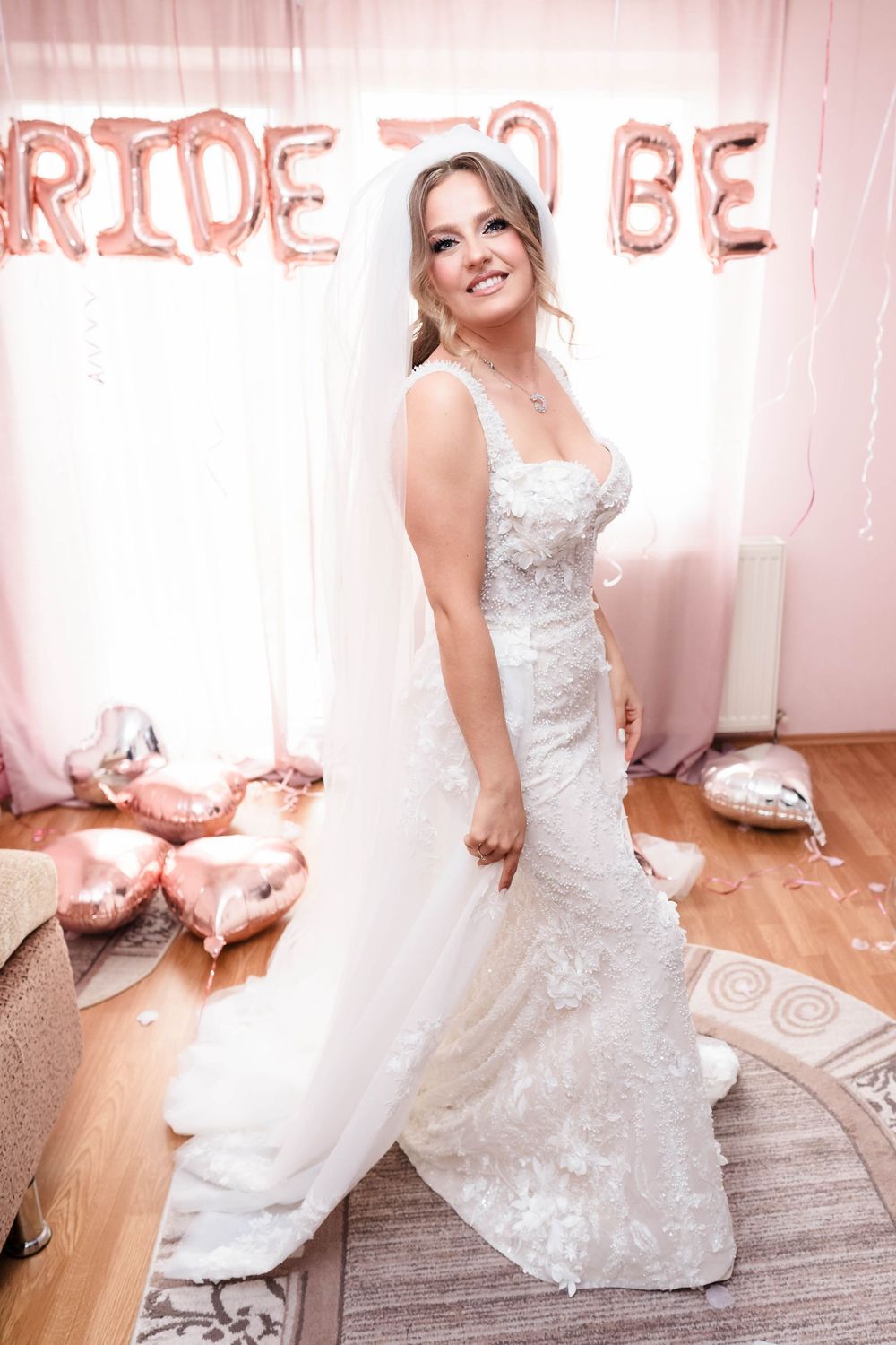 A bride poses in a fitted lace wedding gown against a pink backdrop with 'BRIDE TO BE' balloon letters.