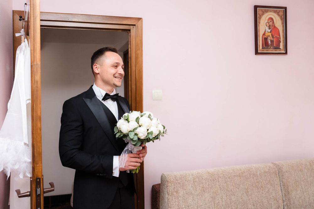 A groom in black tuxedo holds a white bouquet while smiling in a pink-walled room.