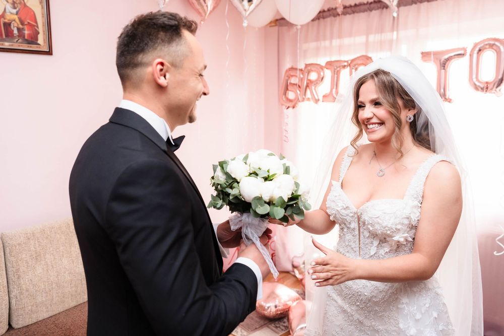 A couple shares a romantic moment exchanging flowers during their wedding day in a pink-decorated room.