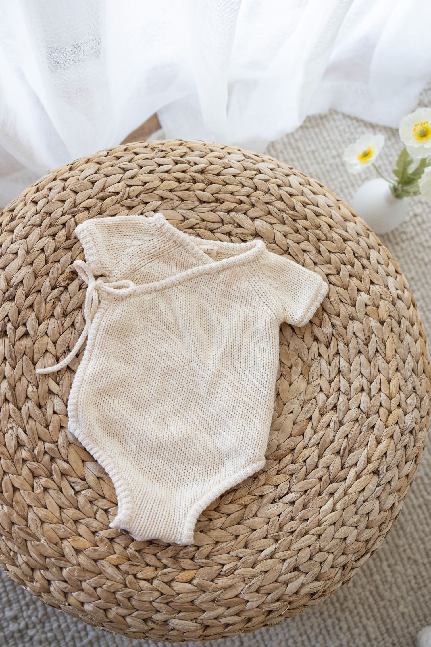 Cream colored knitted baby romper displayed on a round woven straw mat against a white fabric backdrop.