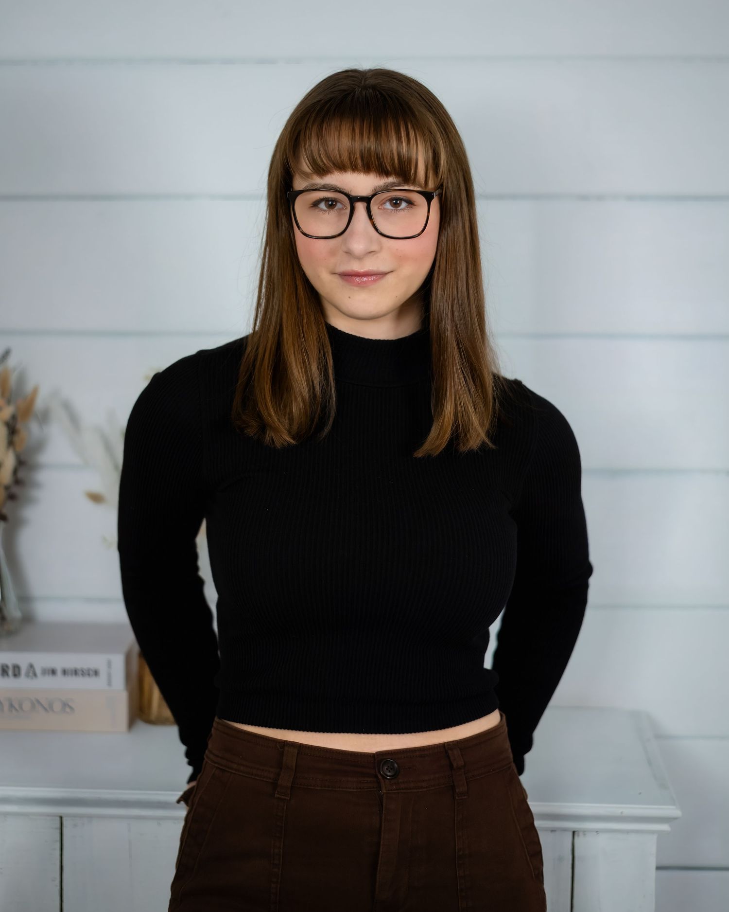 Young person wearing glasses and a black cropped sweater stands indoors against a white wall.