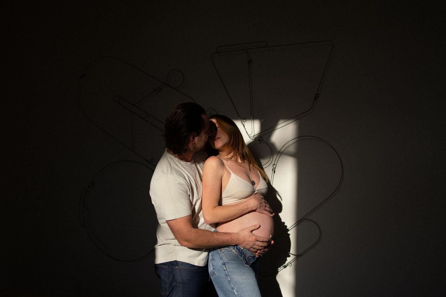 Couple sharing a tender kiss during maternity photo shoot, woman cradling baby bump in soft dramatic lighting.