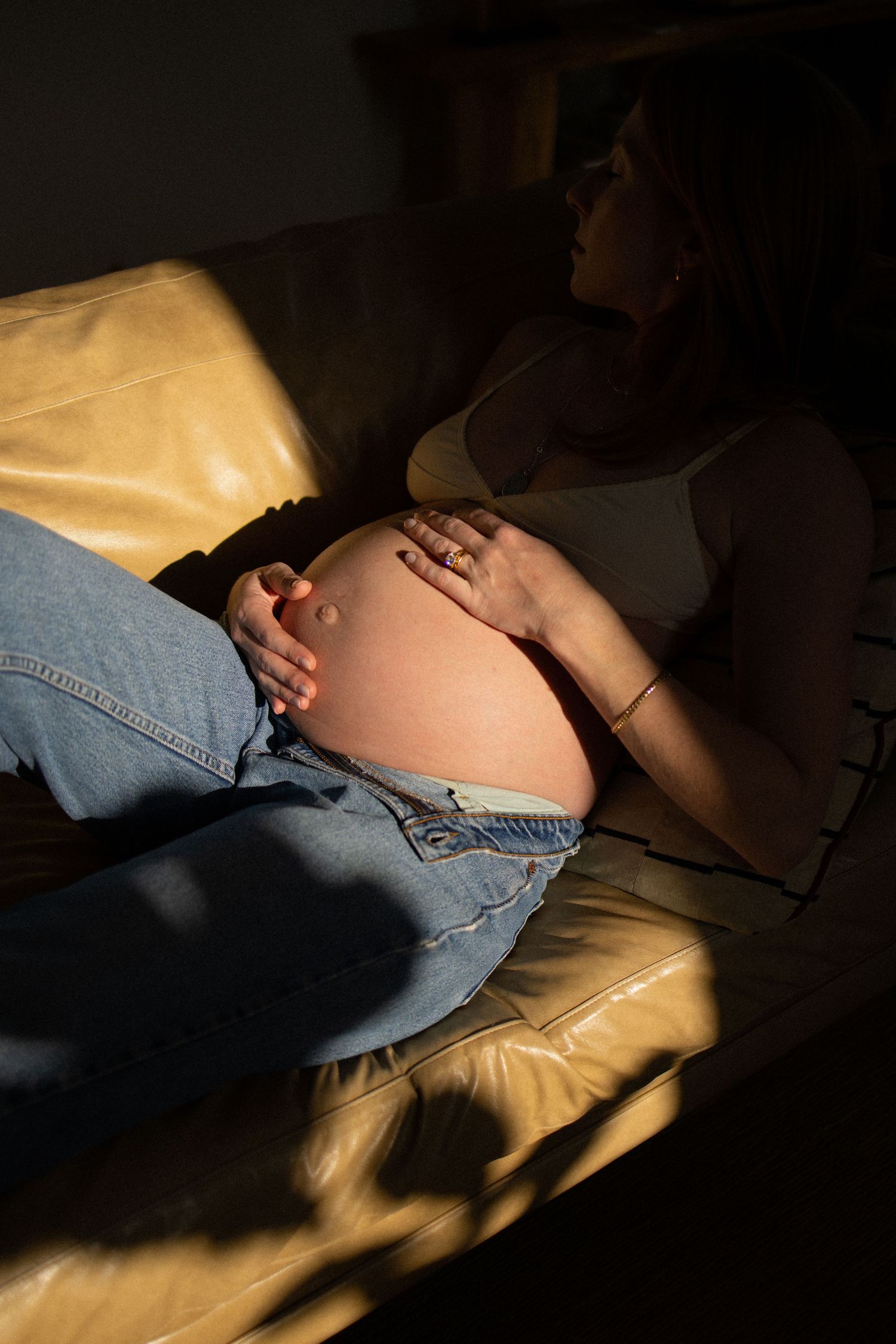 Pregnant woman resting on leather sofa, cradling her bare baby bump, wearing jeans and white top in warm sunlight.
