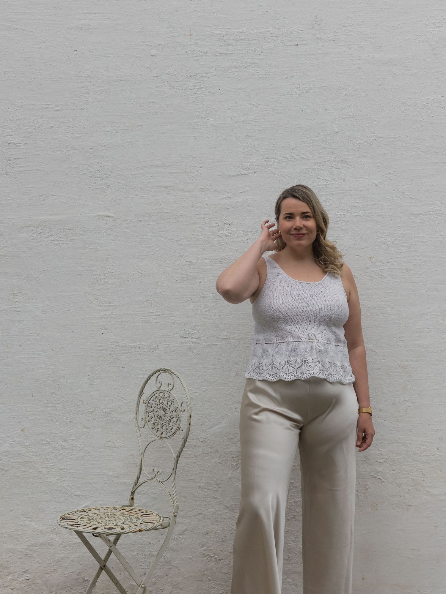 A person in a grey lace-trimmed tank top and beige wide-leg pants stands against a white wall next to a vintage chair.