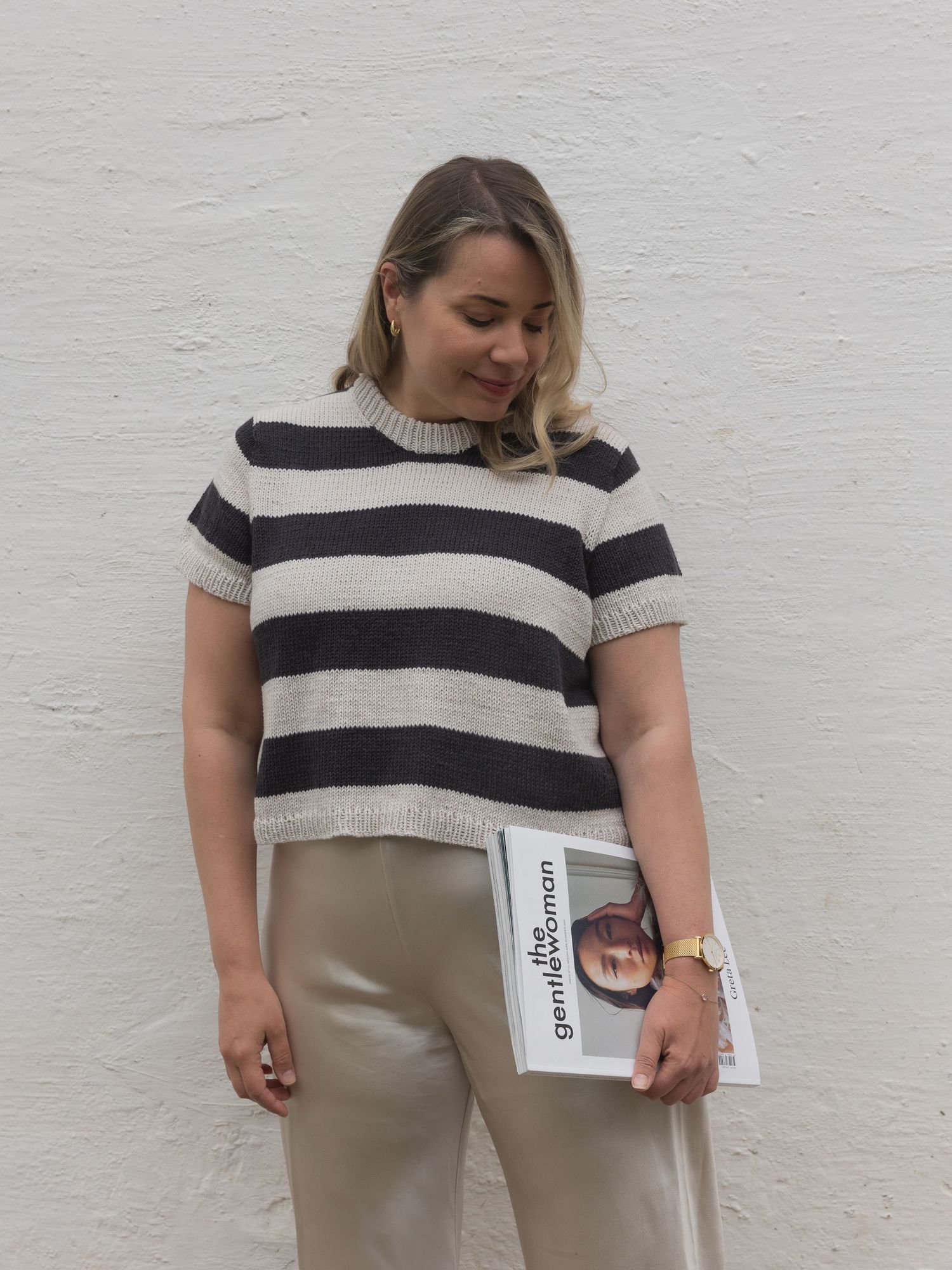 Woman wearing a striped knit short-sleeve sweater holding a book against a white wall.