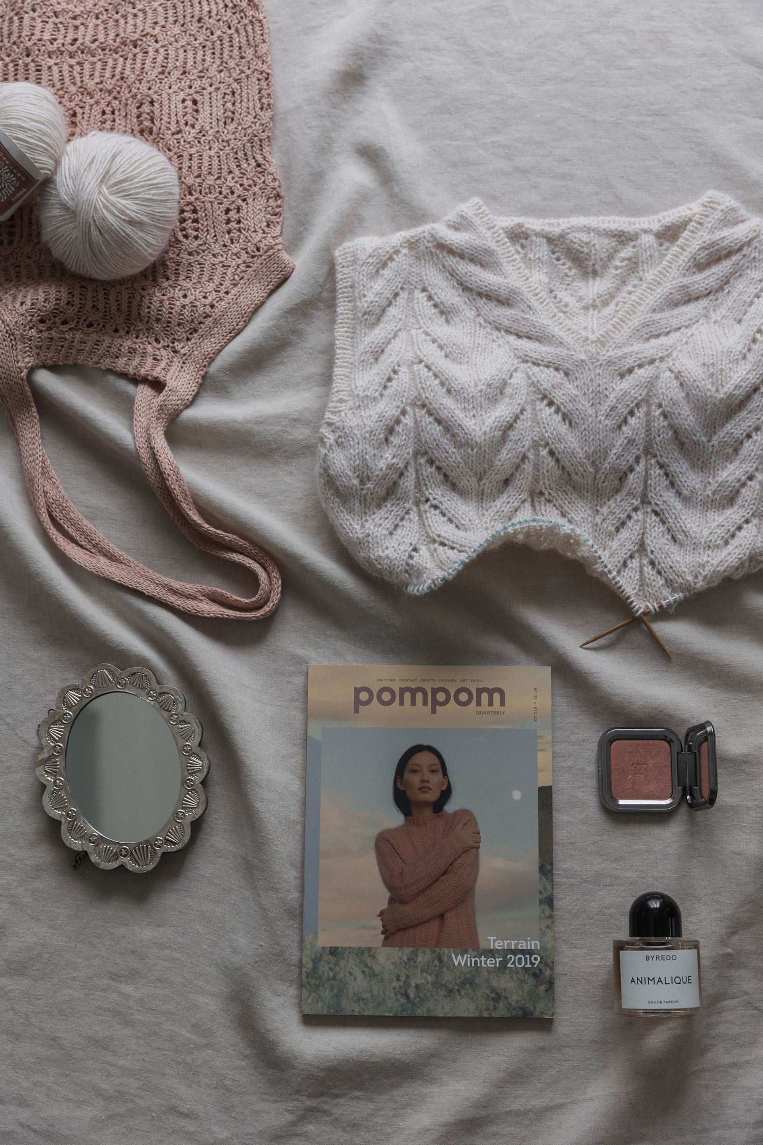 Flatlay of knitting supplies with pink and white yarn, knit samples, decorative mirror, magazine and perfume bottle.