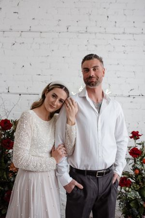 Wedding couple poses against white brick wall with red roses, bride wearing sparkly long sleeve dress.