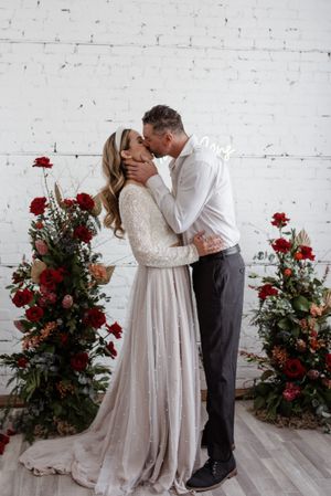 A romantic wedding moment captured between a couple surrounded by red rose arrangements against white brick.