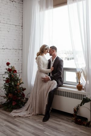 Intimate wedding portrait by window with red rose decorations and white brick wall.