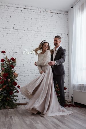 A couple dances joyfully in front of a white brick wall decorated with red roses and a neon sign.