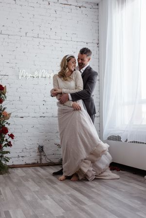 An intimate portrait of a couple with neon 'Mr & Mrs' sign against white brick wall.