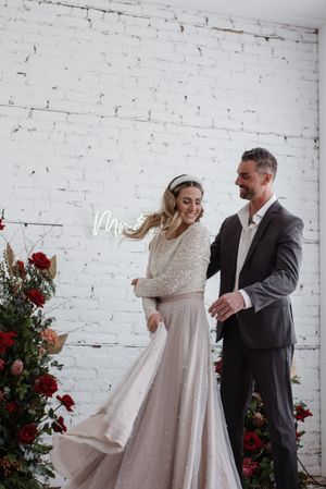Wedding couple share joyful moment dancing together against white brick wall with red rose floral arrangements.
