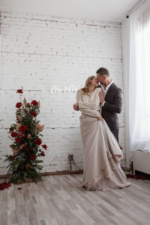 Elegant wedding dance pose against white brick wall with red rose arrangement.