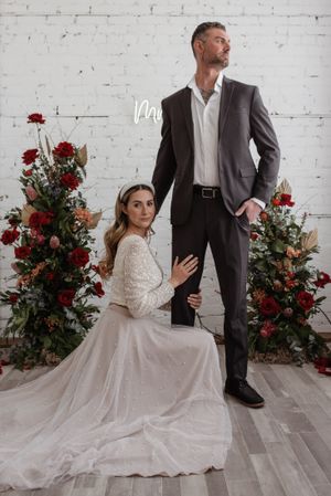 Couple poses by red floral arrangement against white brick wall, bride in lace gown kneeling beside groom in suit.