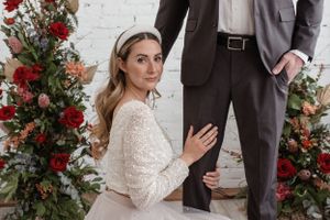 Elegant wedding couple pose with red floral arrangements against white brick wall.