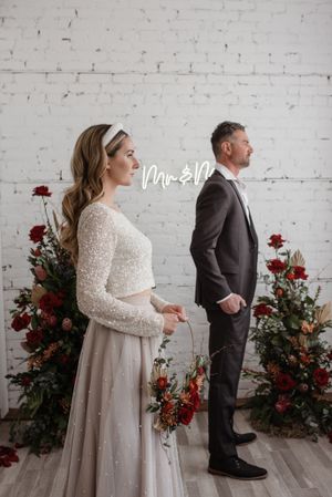 A bride and groom stand back-to-back against a white brick wall with red floral arrangements.
