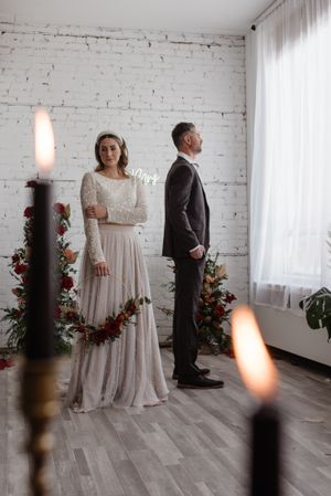 Wedding couple standing separately against white brick wall with floral decorations and candles.