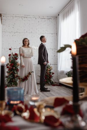 Elegant wedding scene with couple standing by white brick wall adorned with red roses and candles.