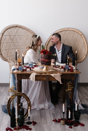 Romantic wedding table setup with rattan peacock chairs, candles, and red floral accents for an intimate celebration.