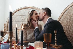 A romantic wedding table setup with candles and champagne against a wicker peacock chair backdrop.