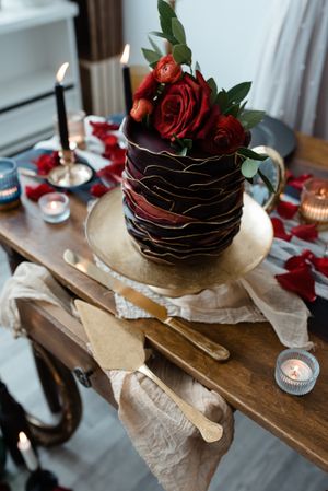 A chocolate wedding cake decorated with red roses sits on a rustic wooden table with elegant place settings.