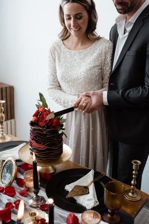 A lovely wedding couple cutting into their elegant dark chocolate tiered cake decorated with red roses.