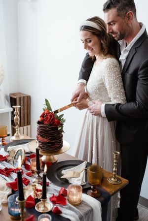 A couple cuts into a chocolate wedding cake topped with red roses on an elegantly decorated table with candles.