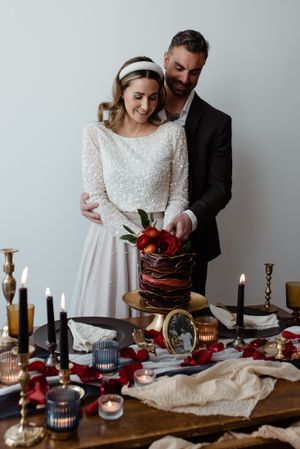 Couple in wedding attire cutting cake together at romantic candlelit table setting with red accents.