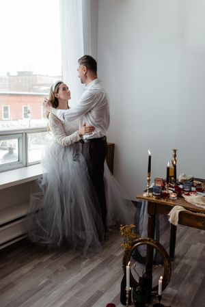 Romantic wedding scene by window with candlelit table setting and flowing tulle dress.